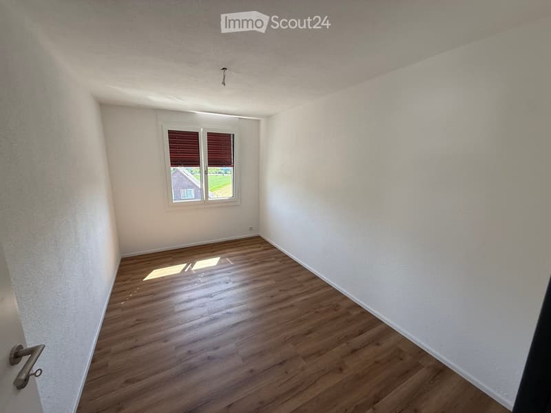 Empty room with wooden flooring and white walls, featuring a window with blinds and a door handle visible.
