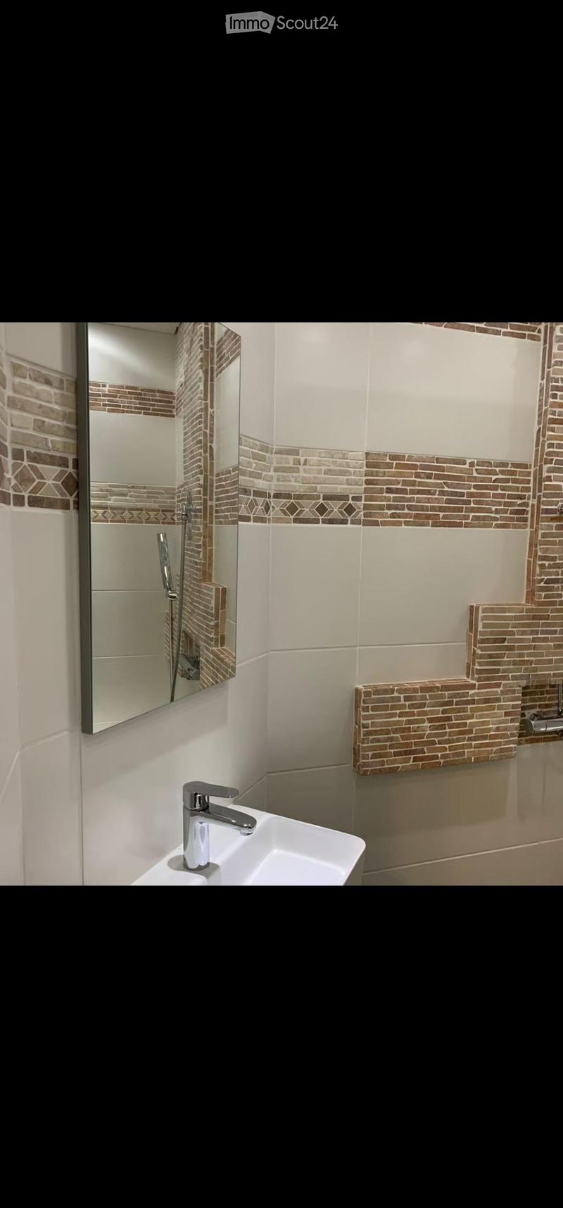 Modern bathroom with white tiles, a decorative brick accent border, a white sink with a chrome faucet, and a large mirror above it. A glass-enclosed shower is visible in the background.