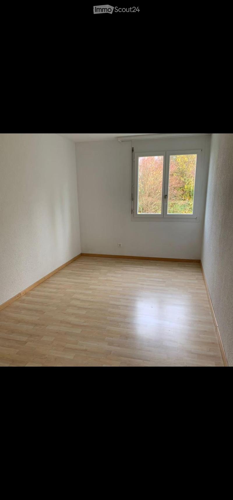 Empty room with light wooden flooring and white walls, featuring a window with a view of greenery outside.
