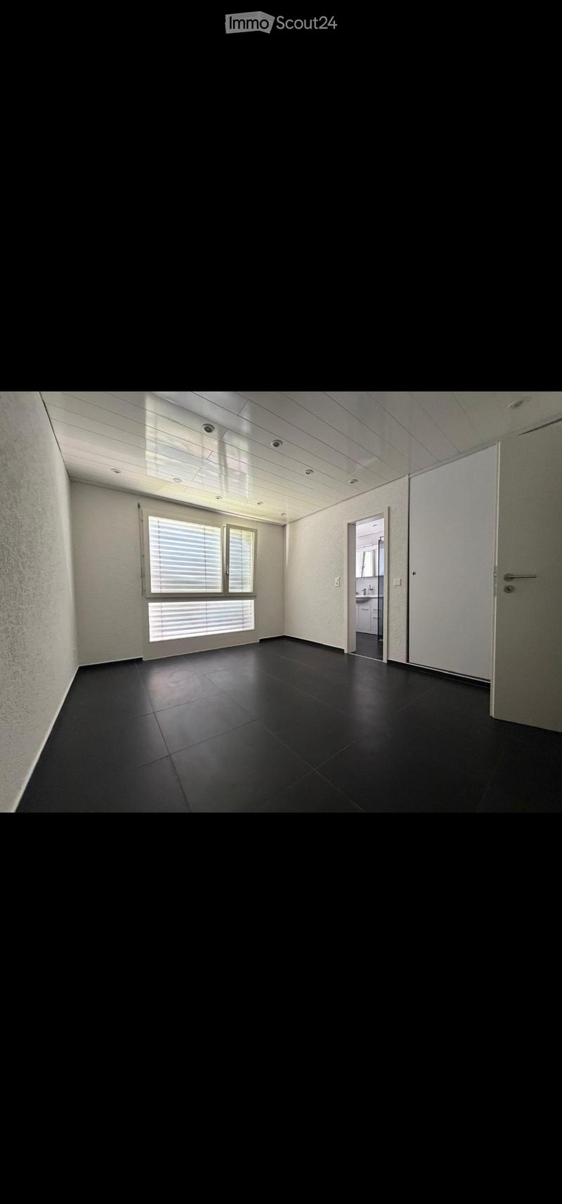 Empty room with black tiled floor, white walls, large window with blinds, and a door leading to a bathroom.