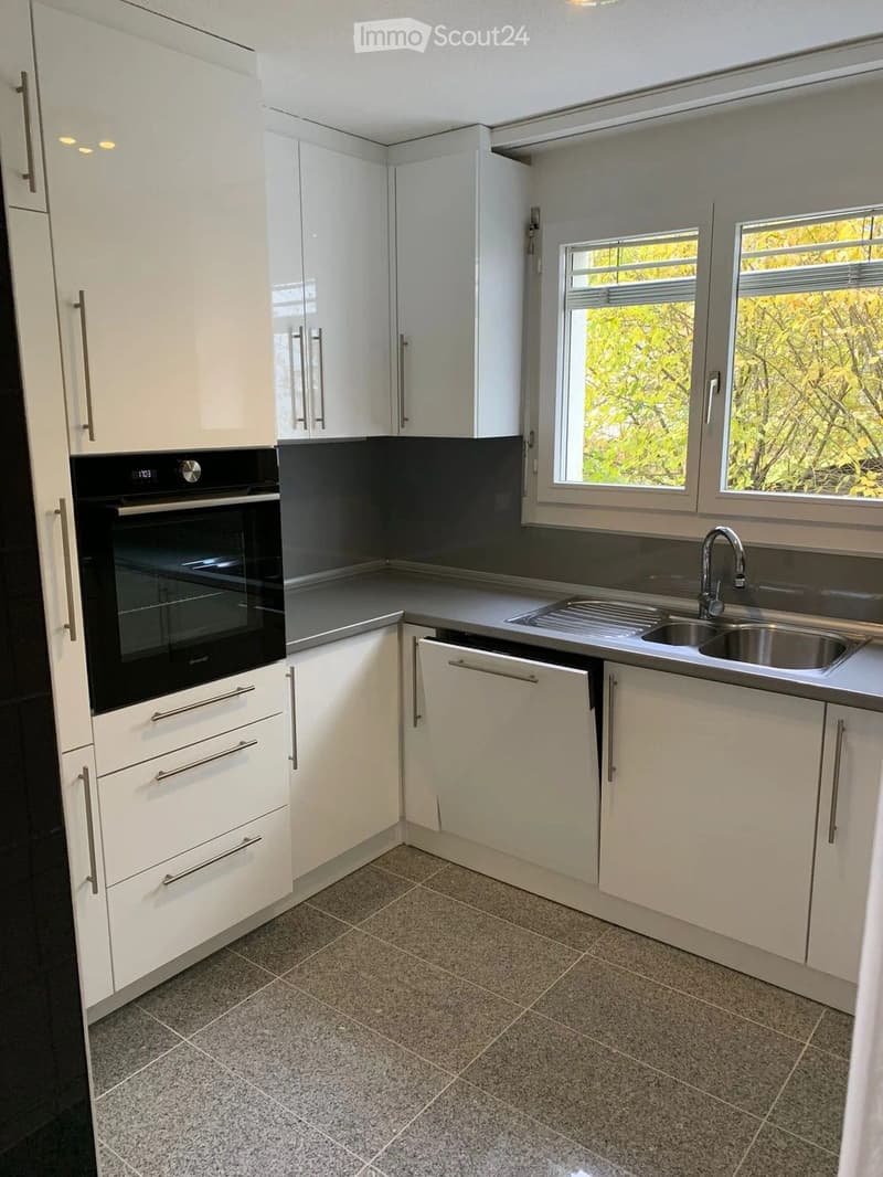 Modern kitchen with white cabinets, stainless steel sink, and built-in oven. Features tiled flooring and large windows with a view of greenery.