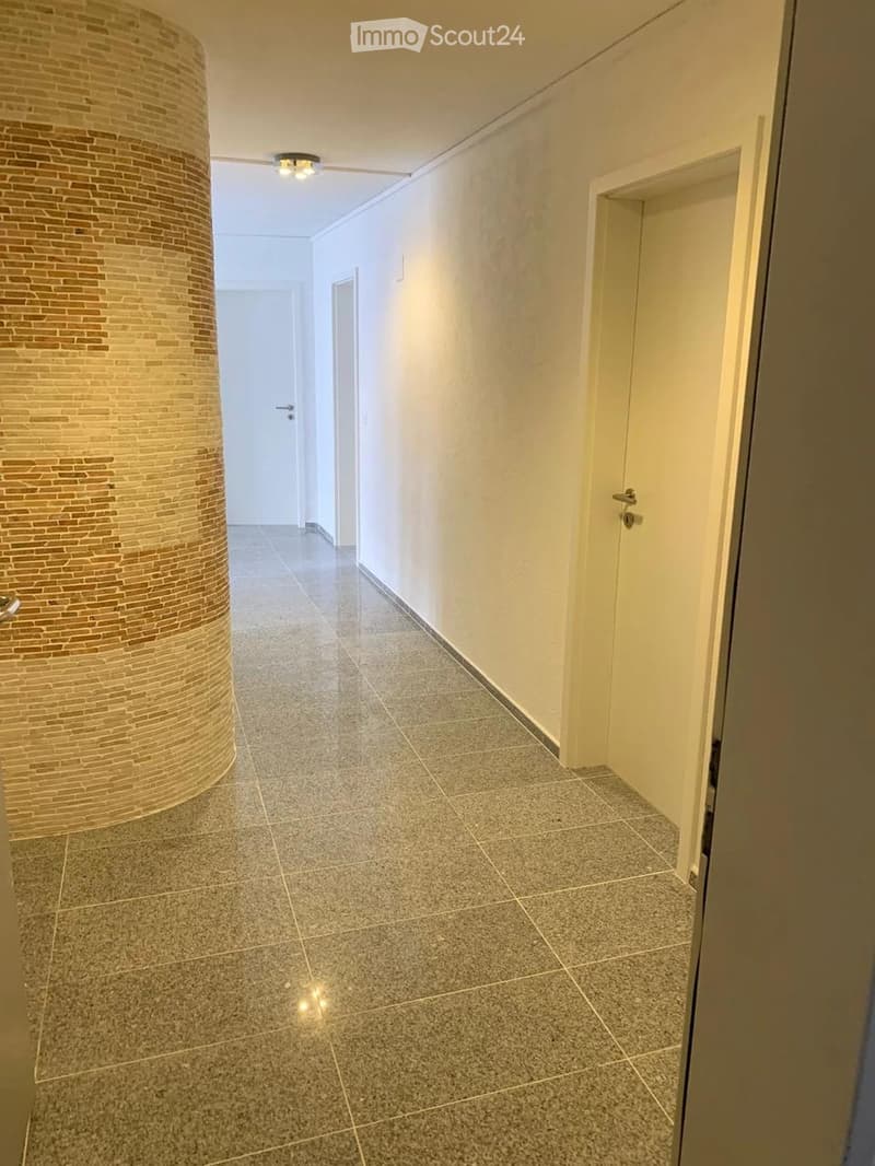 Clean, modern hallway with tiled flooring, white walls, and recessed lighting. Features a decorative brick pillar on the left and several doors along the right side.