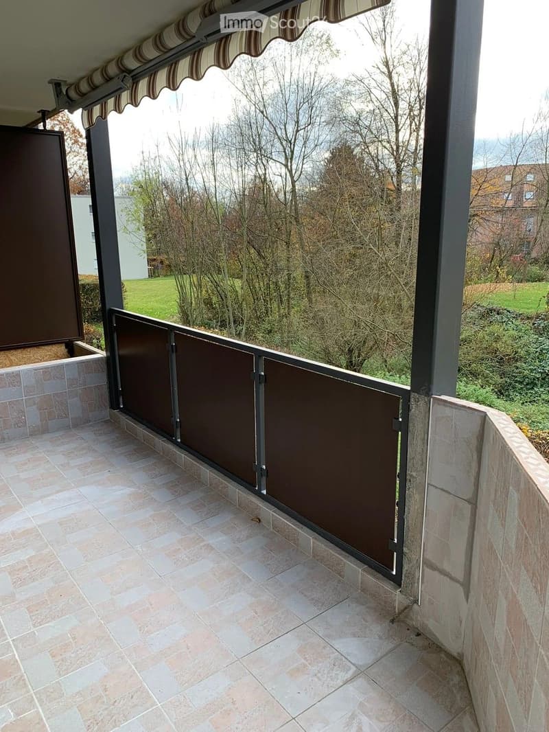 Balcony with tiled floor, brown privacy screen, and striped awning, overlooking a green landscape with trees and distant houses.