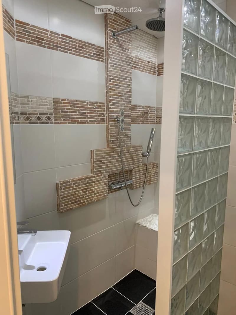 Modern bathroom with white sink, dark tiled floor, and glass block wall. Features a shower area with a handheld showerhead and a built-in seat.