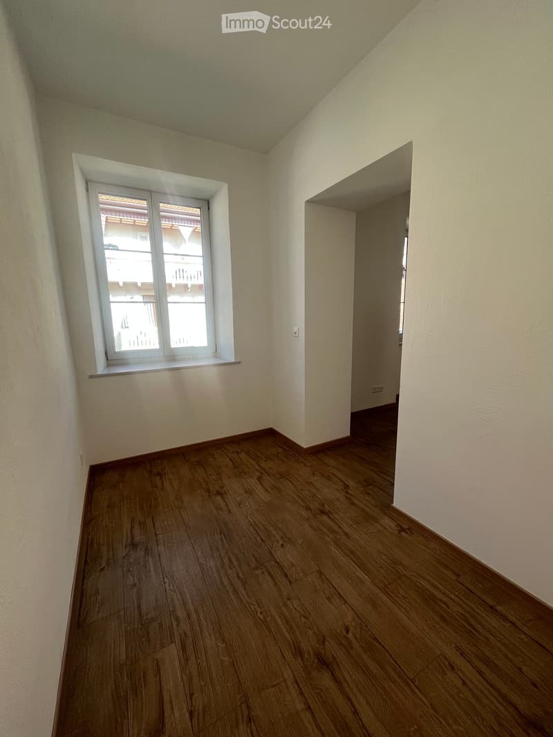 Empty room with wooden flooring, white walls, and a window with a view of a balcony and neighboring building.