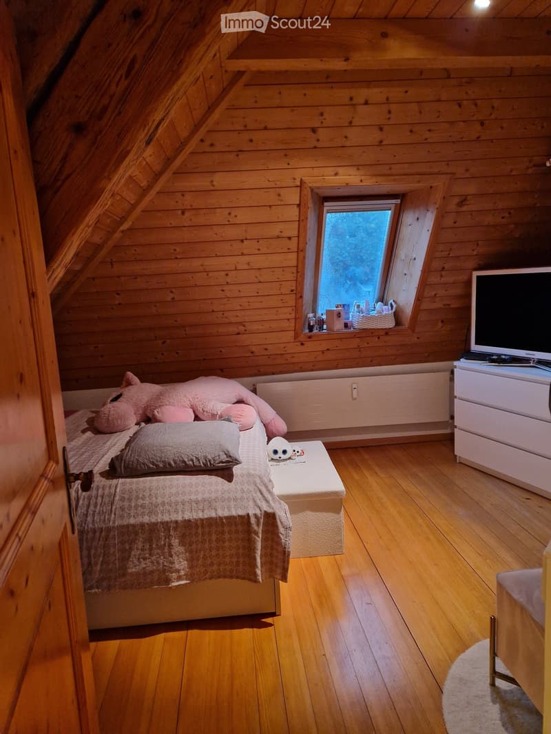 Cozy attic bedroom with wooden walls and floors, featuring a bed with a pink stuffed animal, a small table with a television, and a window with a view.