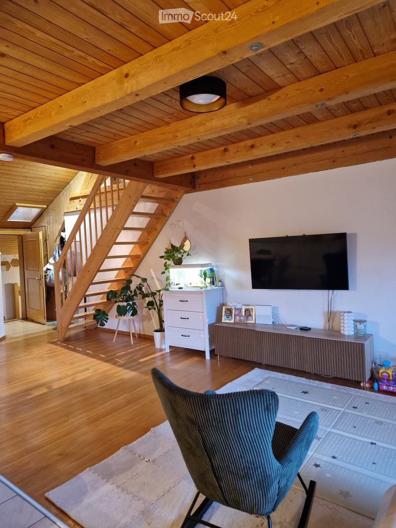 Living room with wooden floors, exposed wooden beams, a staircase, a television mounted on the wall, a grey armchair, a small table with plants, and a cabinet with picture frames.