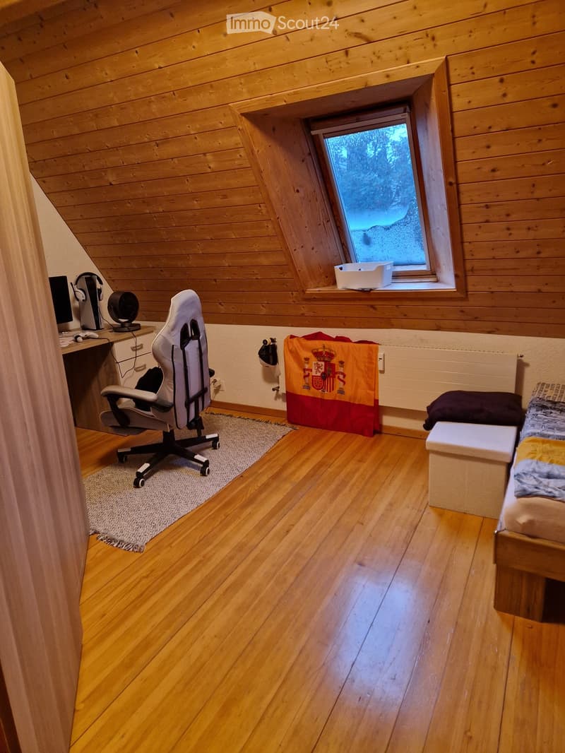 Cozy attic room with wooden walls and floors, featuring a bed, desk with chair, and a window with a view.