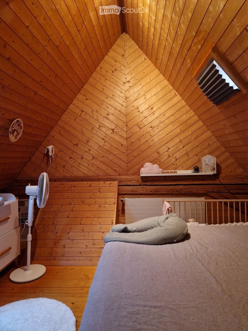 Cozy bedroom with wooden walls and slanted ceiling, featuring a bed with a grey blanket, a fan, a nightstand, and a shelf with decorative items.