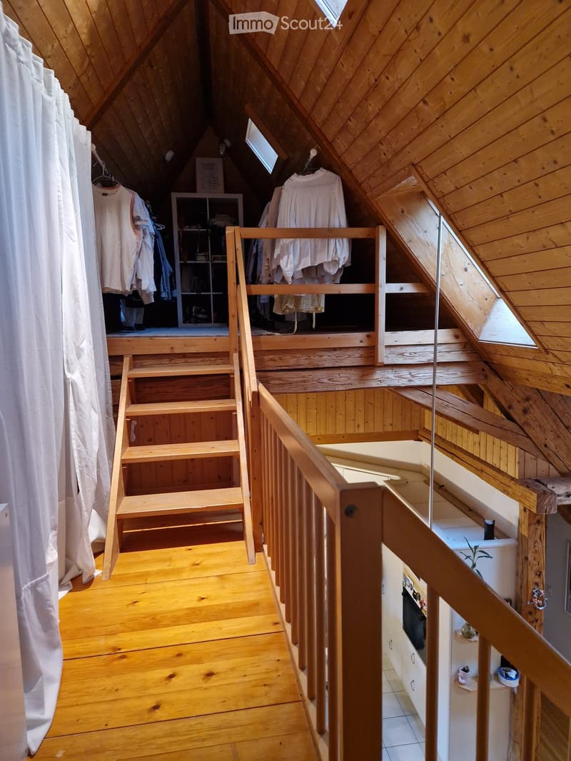 Wooden staircase leading to an attic space with slanted wooden ceilings and built-in storage shelves. The lower level features wooden flooring and a small white cabinet with a plant on top.