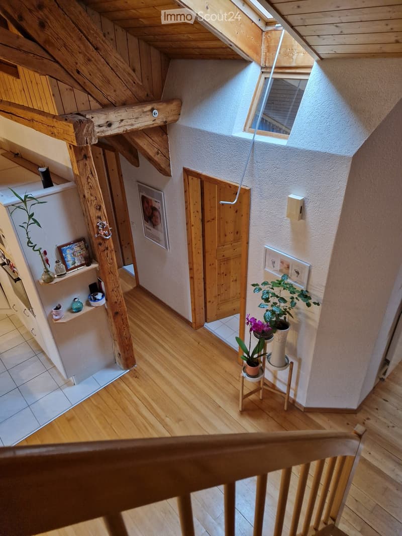 Wooden staircase with slanted ceiling, exposed wooden beams, small window, potted plants, and framed pictures on walls.