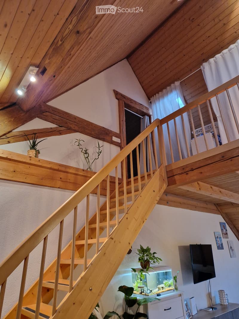 A cozy living space with a wooden staircase leading to an upper level, a fish tank on a cabinet, a television mounted on the wall, and several potted plants adding greenery to the room.