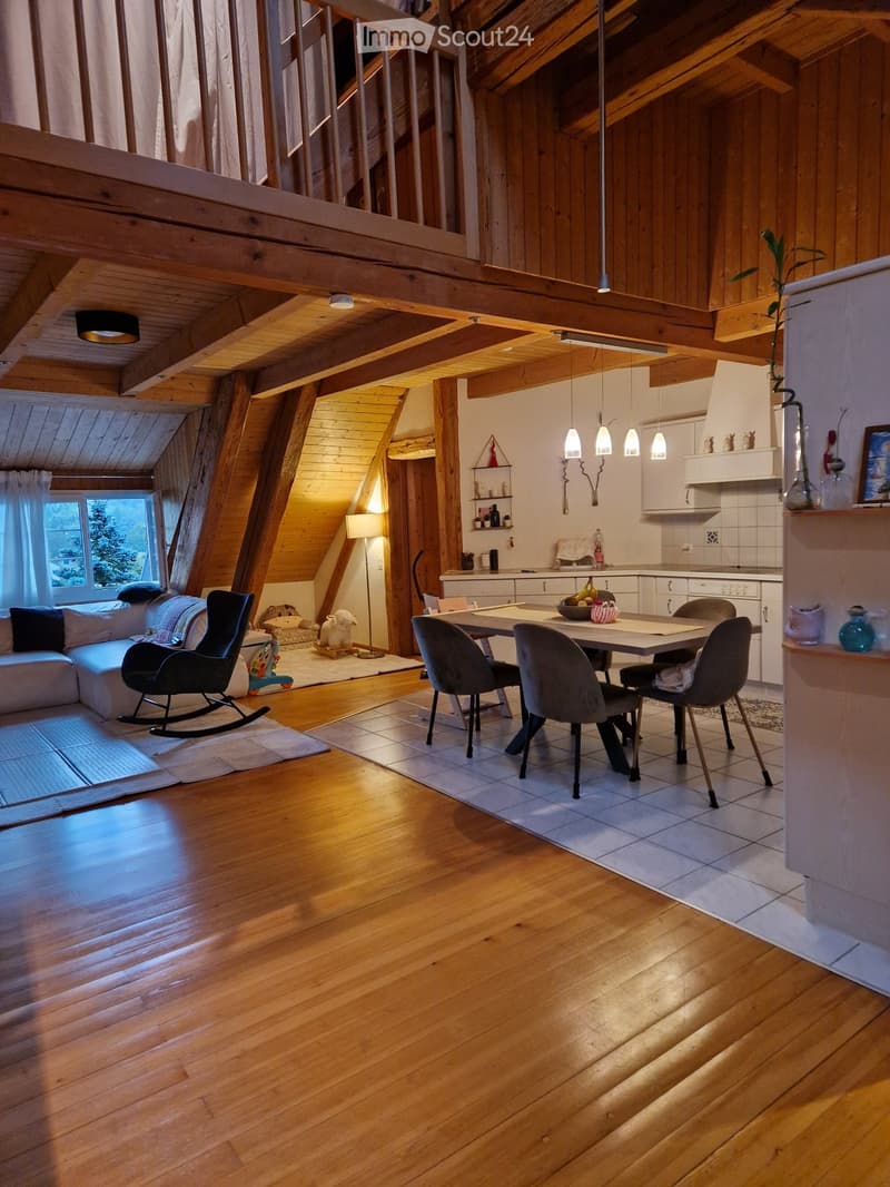 Open-plan living and dining area with wooden beams, hardwood floors, a sofa, rocking chair, dining table with chairs, and a small kitchen area with cabinets and shelves.