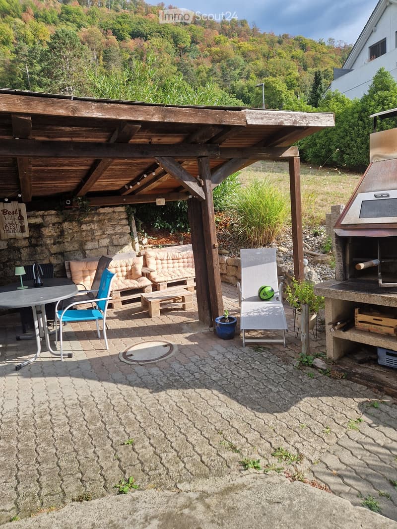 Outdoor patio with stone flooring, wooden pergola, seating area including a sofa, chairs, and a table, along with a barbecue and potted plants.