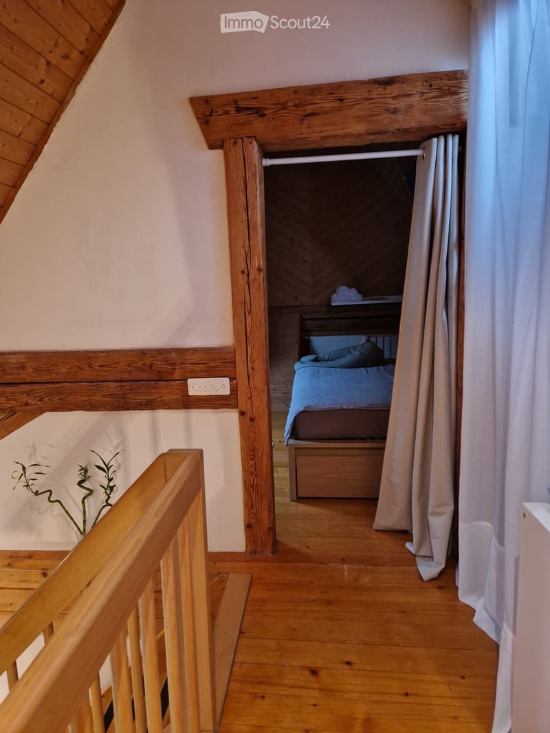 Cozy attic bedroom with wooden bed, blue bedding, and a curtained window. Wooden floor and slanted ceiling beams.