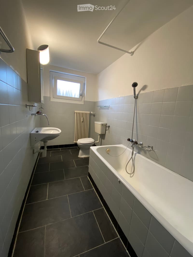 Bathroom with a bathtub, toilet, sink, radiator, and window. Dark tiled floor and light grey tiled walls.