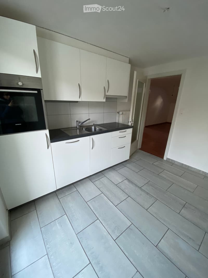 Modern kitchen with white cabinets, stainless steel sink, and oven. Grey tiled floor and black countertop.