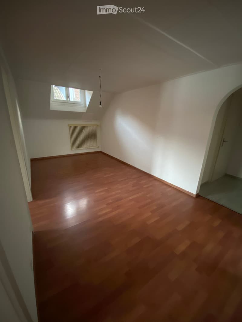 Empty attic room with wooden flooring, white walls, a small window, and a radiator.