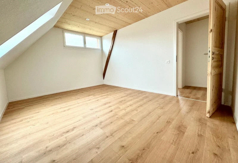 Empty attic room with wooden flooring, white walls, skylight window, and exposed wooden ceiling beams.