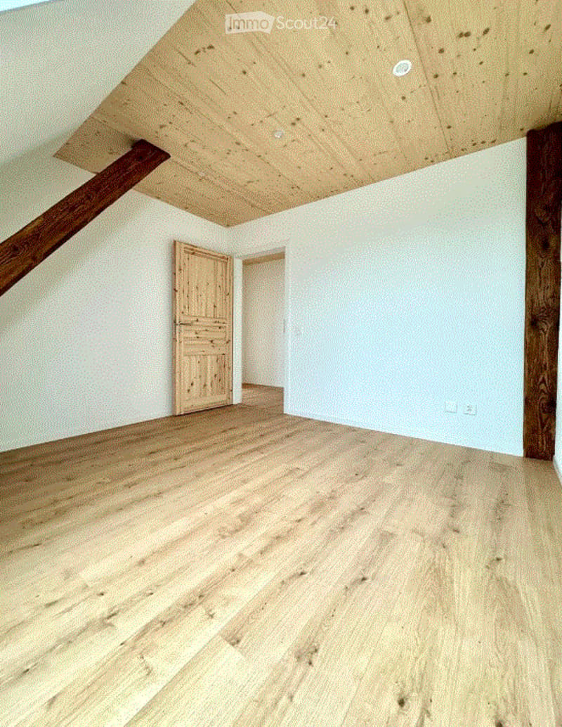 Empty attic room with wooden floors, white walls, and exposed wooden beams. Features a wooden door and a small open doorway leading to another space.