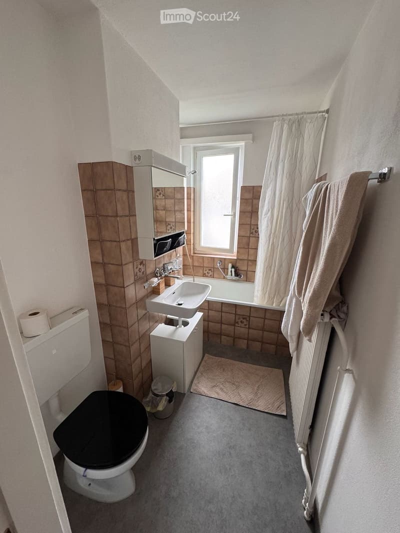 Bathroom with a toilet, sink, bathtub, mirror, and a towel hanging on a rack. The room features tiled walls, a window, and a radiator.