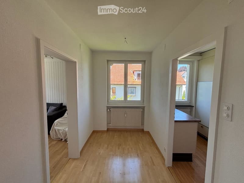 A small, unfurnished living room with wooden flooring, white walls, and large windows showing a view of neighboring houses. A sofa and a table are partially visible.