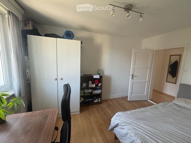 Cozy bedroom with wooden flooring, a bed with striped bedding, a chair, a small table, a wardrobe, a shelf with books and items, and a plant near the window.