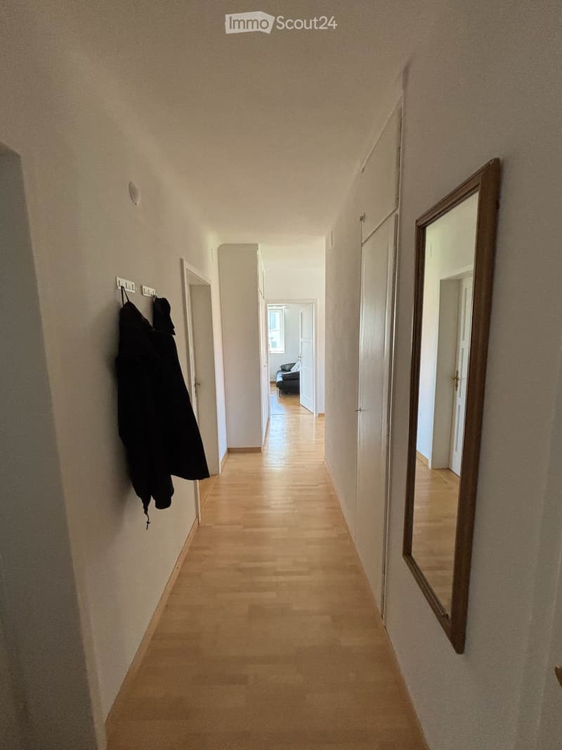 A long, narrow hallway with light wooden flooring, beige walls, and a large mirror on the right side. A coat hangs on the left wall, and a glimpse of a room with a sofa is visible at the end.