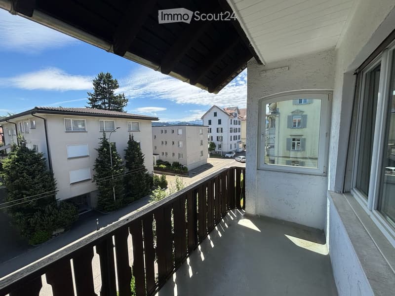 Balcony with wooden railing, overlooking a street with buildings and trees under a clear blue sky.