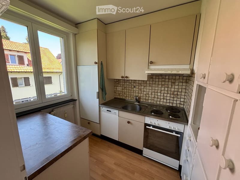 Bright kitchen with white cabinets, a refrigerator, dishwasher, oven, sink, and a countertop. The room has wooden flooring and large windows offering a view of a neighboring building.