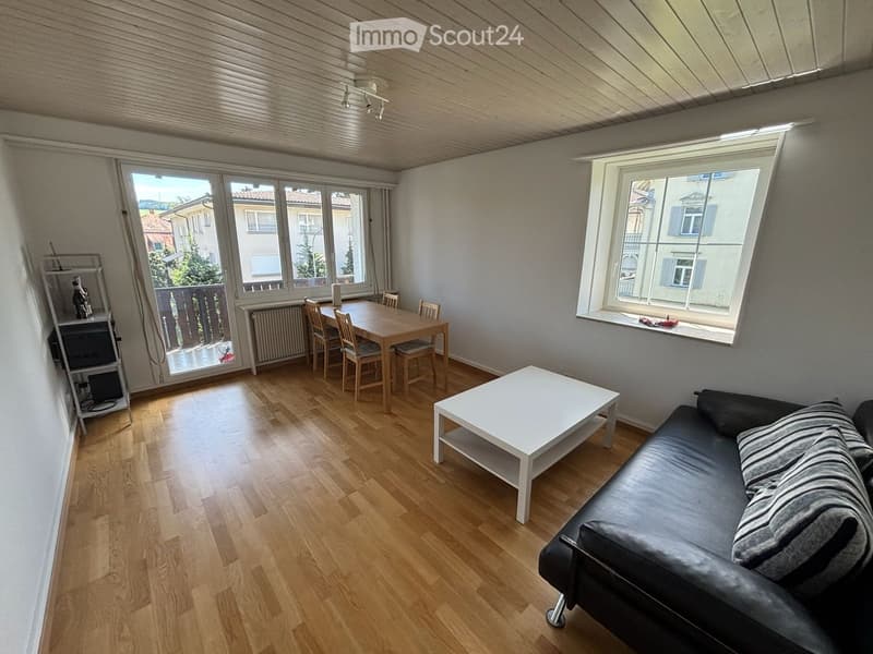Bright living room with wooden flooring, large windows, a black leather sofa, a white coffee table, a dining table with chairs, and a balcony view.