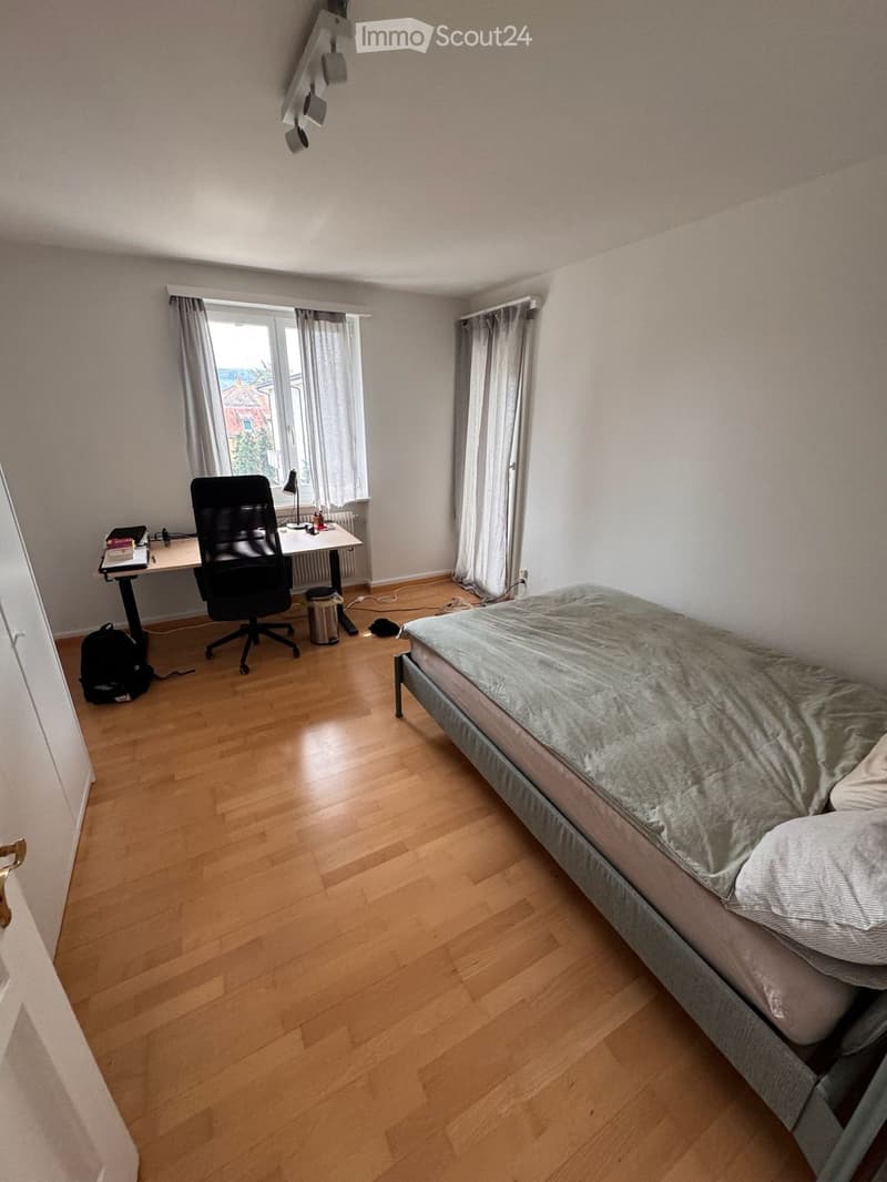 A furnished bedroom with a bed, desk, chair, and lamp. The room features wooden flooring and large windows with curtains.