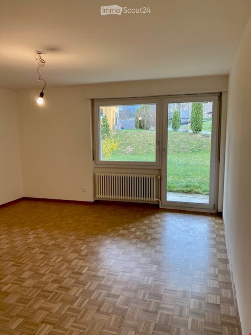 Empty room with wooden parquet flooring, white walls, a radiator, and large sliding glass doors leading to a green outdoor area.