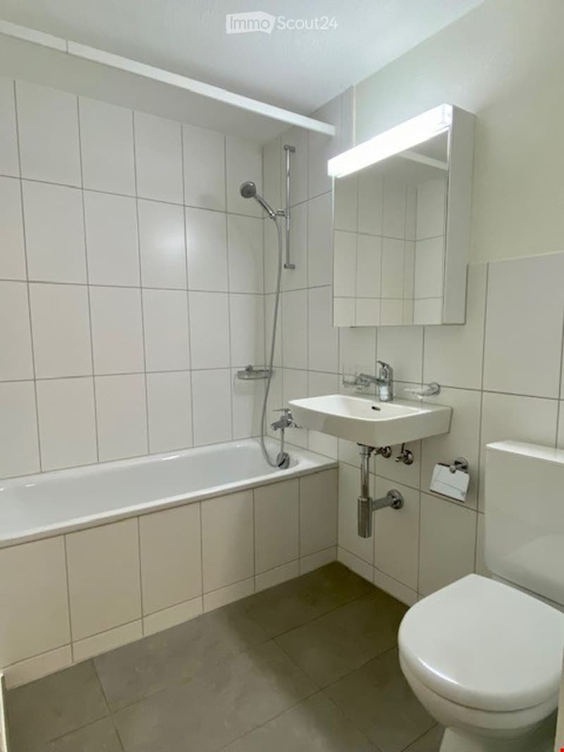 Modern bathroom with white tiled walls and floor, featuring a bathtub, toilet, sink, and a mirror with built-in lighting above the sink.