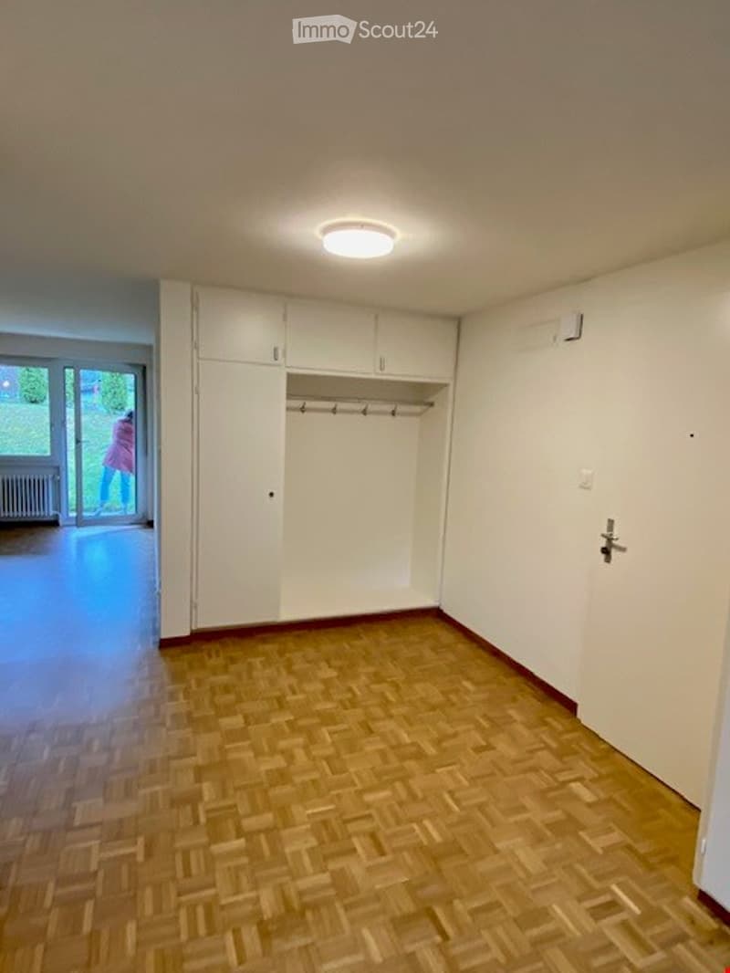 Empty room with wooden parquet flooring, white walls, built-in white closet with hanging rod, and a glass door leading to a balcony or outdoor area.