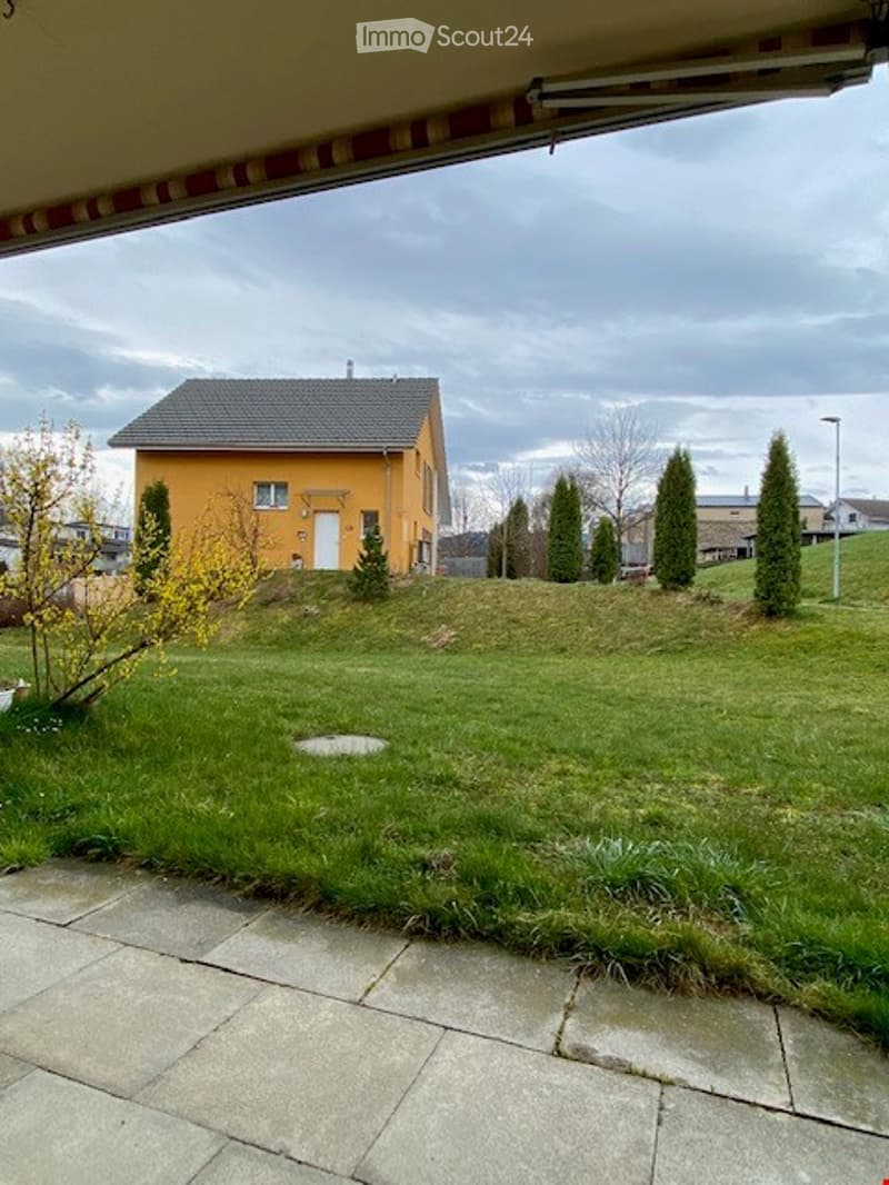 A well-maintained garden with a yellow house in the background, featuring a lawn, small trees, and a paved patio area.