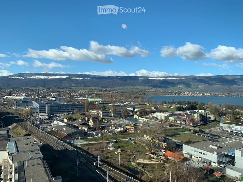 Aerial view of a town with buildings, railway tracks, and a body of water in the background, surrounded by mountains.