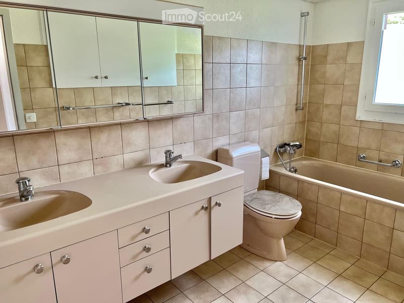 Modern bathroom with beige tiles, double sink vanity, toilet, bathtub, and large mirror with cabinets above.