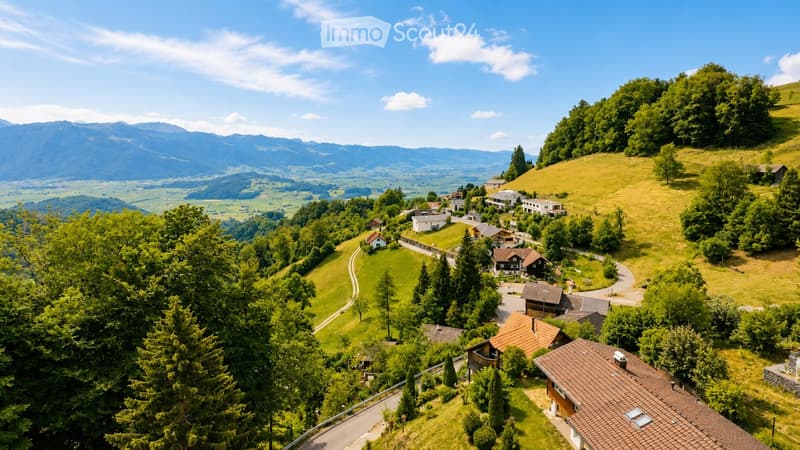 Aerial view of a rural village nestled in a lush green valley surrounded by mountains. The village features scattered houses with various roof styles, surrounded by dense forests and open fields.