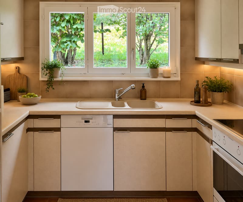 Modern kitchen with light-colored cabinets, a double sink, a dishwasher, and an oven. The countertop features a bowl of fruit, a bottle, a candle, and potted plants. A window with a view of greenery is above the sink.