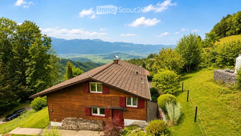 A wooden house with a brown tiled roof, surrounded by lush greenery and a scenic mountain view in the background.