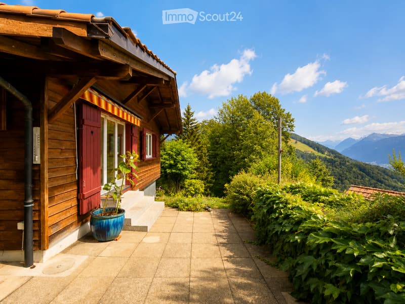 A wooden house with a red door and a blue planter on the patio, surrounded by lush greenery and a scenic mountain view.