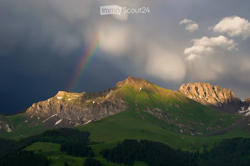 A scenic mountain landscape with a prominent peak, lush green valleys, scattered trees, and a rainbow visible in the sky.