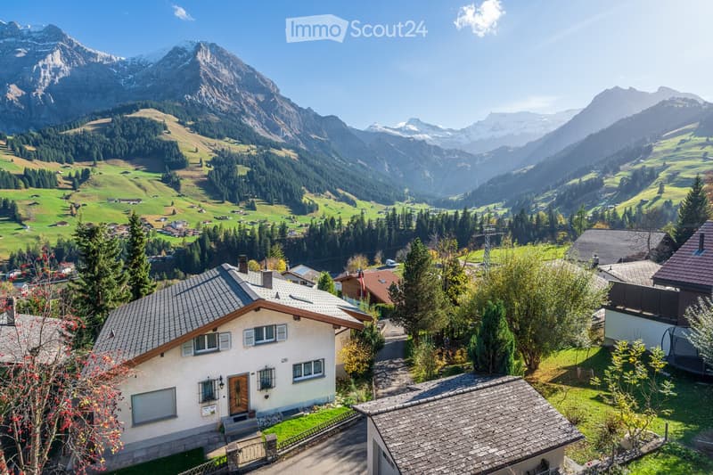 A picturesque view of a Swiss village nestled in a lush green valley surrounded by snow-capped mountains. The village features traditional houses with pitched roofs and wooden facades, surrounded by trees and greenery.