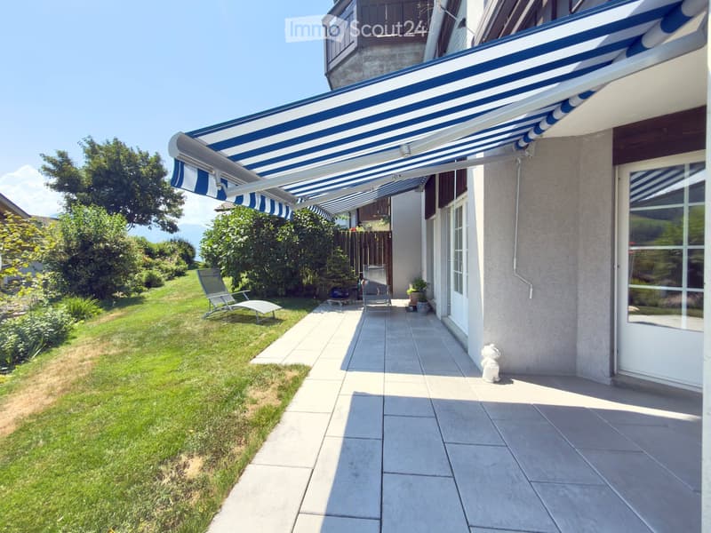 A covered patio area with a blue and white striped awning, tiled floor, two chairs, and a small table, adjacent to a well-maintained lawn and garden.