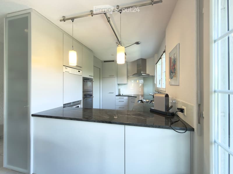 Modern kitchen with white cabinets, dark granite countertop, built-in oven, range hood, and pendant lights. Coffee maker on the counter.