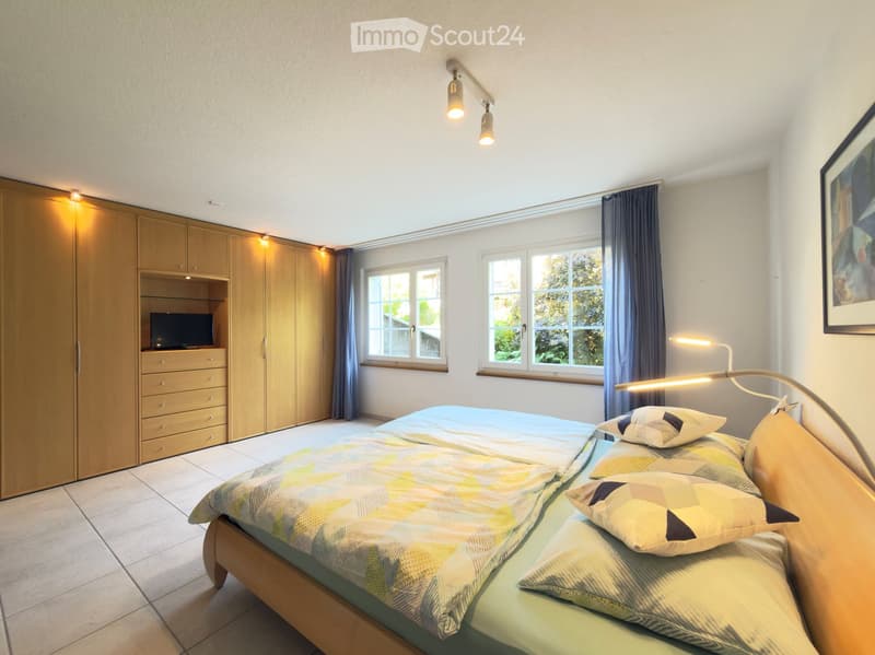 Bright bedroom with a large bed, colorful bedding, built-in wardrobe, TV, and large windows with curtains.