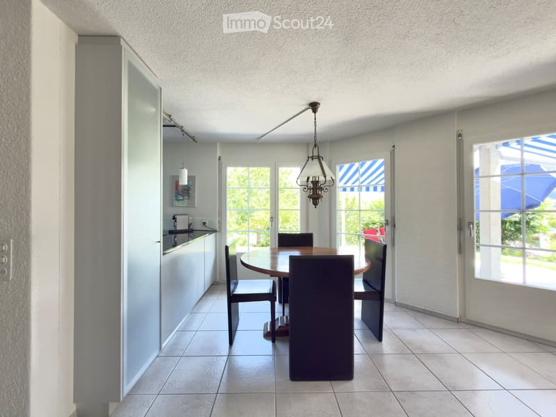 Bright, open dining area with a round wooden dining table, four chairs, and a bench, connected to a small kitchen area with a counter and sink.