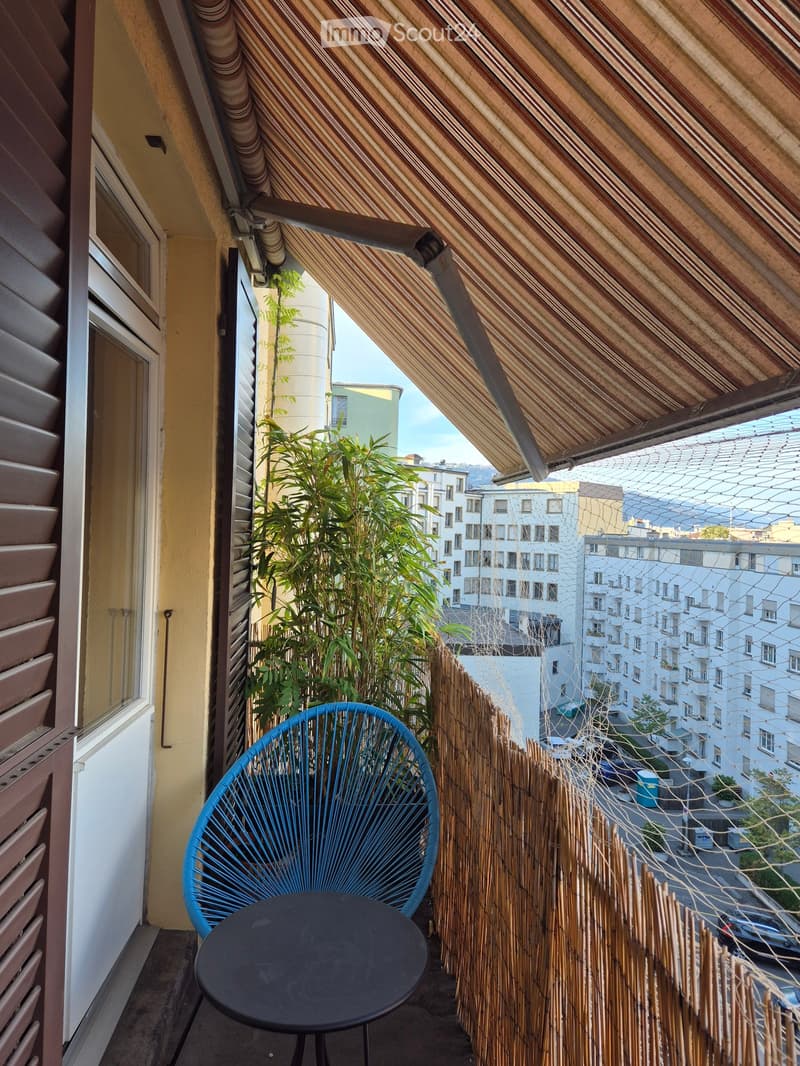 Balcony with a blue wire chair, black round table, bamboo screen, and potted plants, overlooking a cityscape.