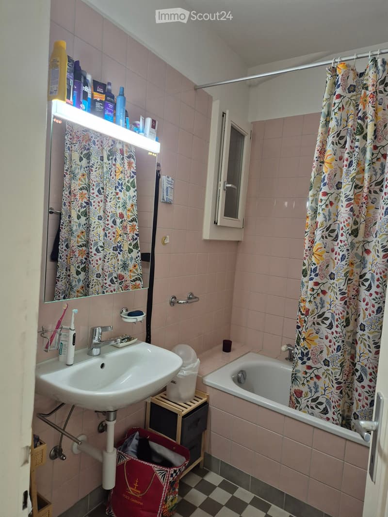 Pink-tiled bathroom with a sink, mirror, bathtub, and shower curtain. Includes a small window, a wooden shelf, and various toiletries.